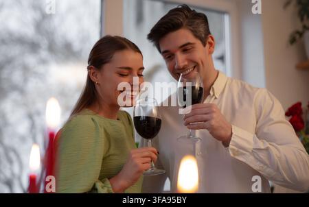 Giovane coppia sorridente solleva bicchieri di vino rosso accanto a candele calde durante una romantica celebrazione di San Valentino a casa. Foto Stock