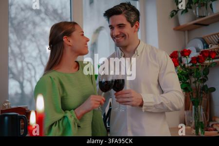 Una giovane coppia felice tiene bicchieri di vino rosso e si sorride l'un l'altro accanto a rose rosse e candele accese in una cucina di casa. Foto Stock