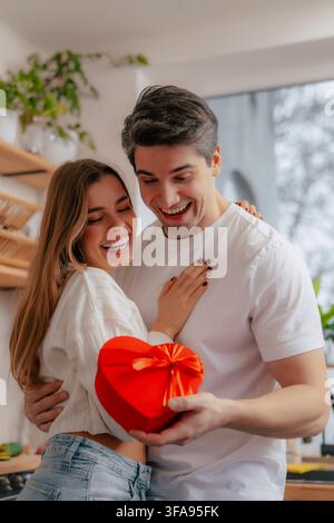 Ritratto verticale in stile lifestyle di una giovane coppia gioiosa che si abbraccia e guarda una scatola regalo rossa a forma di cuore in una cucina luminosa. Foto Stock