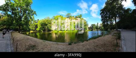 Vista panoramica di un lago sereno del parco circondato da alberi verdi rigogliosi, con una grotta rocciosa sullo sfondo sotto un cielo blu. Foto Stock