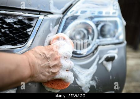 Primo piano di una mano che lava il faro di un'auto con una spugna arancione e schiuma di sapone, pulizia del veicolo, dettagliazione auto, manutenzione e concetto di lavaggio manuale dell'auto. Foto Stock