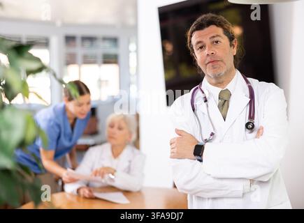 un collega medico maschio in piedi nella sala riunioni e i dipendenti parlano dietro di lui al tavolo Foto Stock