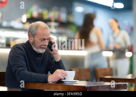 un uomo anziano beve caffè e tiene in mano un telefono cellulare Foto Stock