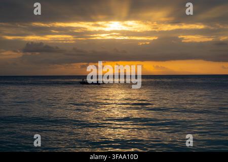 Tramonto dorato sull'oceano con una barca in silhouette, nuvole stratificate e riflessi caldi sull'acqua calma del mare. Foto Stock