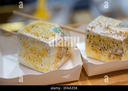 Due grandi marshmallow a forma di cubo su spiedini di bambù, torrefatti e caramellizzati con cristalli di zucchero fini e briciole, serviti in vassoi di carta bianca a un Foto Stock