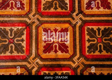 Vista ravvicinata di piastrelle decorative invecchiate con motivi geometrici e floreali ripetuti in rosso, arancione e marrone, che mostrano graffi, usura e stile tradizionale Foto Stock