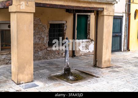 Fontana storica in metallo con acqua corrente si trova su un lastricato di pietra accanto a un edificio scolorito con mattoni a vista, colonne e persiane verdi. Foto Stock