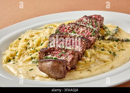 Bistecca di manzo grigliata affettata servita su risotto cremoso con uovo in camicia e guarnizione di cipolla verde su un piatto bianco su uno sfondo beige caldo, casua di alto livello Foto Stock