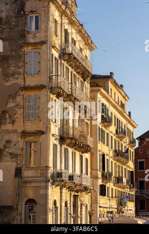 Corfù, Grecia, 10 luglio 2026: Antichi edifici tradizionali con balconi fiancheggiano una strada immersa nel caldo sole del tardo pomeriggio Foto Stock