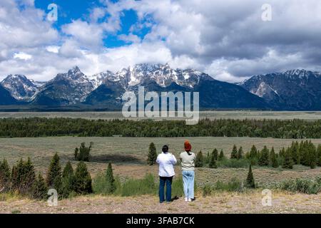 Vista panoramica di due visitatori che ammirano la catena dei Teton innevata sotto nuvole drammatiche da un punto panoramico nel Parco Nazionale di Grand Teton, Wyoming Foto Stock