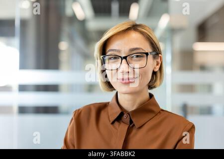 Imprenditrice professionale con occhiali e camicia marrone, in piedi con sicurezza in un ambiente d'ufficio moderno, che trasmette professionalità e successo aziendale Foto Stock