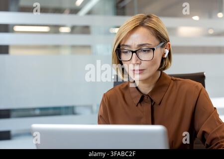 Donna d'affari con auricolari senza fili e occhiali, concentrata sul lavoro utilizzando un laptop in un moderno ufficio aziendale, incarnando concentrazione e produttività Foto Stock