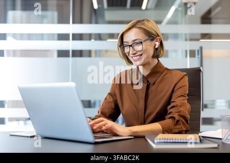 Donna professionista sorridente, con occhiali e auricolari senza fili mentre digita su un laptop, attivamente impegnata nella comunicazione online e nei compiti di lavoro dal suo spazio di lavoro contemporaneo Foto Stock