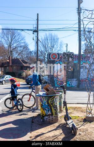 Atlanta, Georgia, Stati Uniti - 18 gennaio 2021: Vista di un uomo e un bambino in bicicletta in attesa a un angolo di strada con un bidone della spazzatura coperto di graffiti Foto Stock