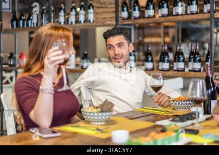 Giovani adulti che condividono vino rosso durante una conversazione intima al ristorante. Una coppia millennial o Gen Z si gode una degustazione rilassata in un'accogliente wine bar. Foto Stock