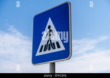 Un cartello quadrato blu con un triangolo bianco che indica un passaggio pedonale è montato su un palo di metallo contro un cielo sereno Foto Stock