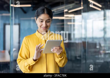 Una giovane donna indiana è in piedi in un ufficio e usa un tablet sorridendo. Foto Stock
