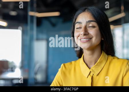 Foto ravvicinata di una giovane indiana sognante in ufficio, seduta comodamente con gli occhi chiusi e che riposa Foto Stock