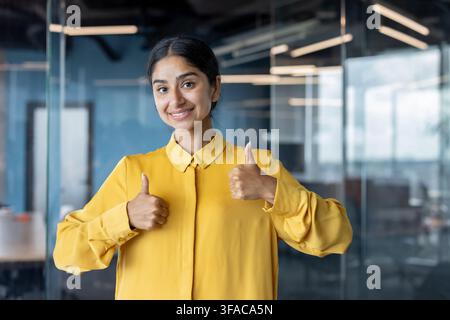 Ritratto di una giovane donna indiana in piedi in ufficio che mostra alla telecamera un segno di successo con le dita. Foto Stock