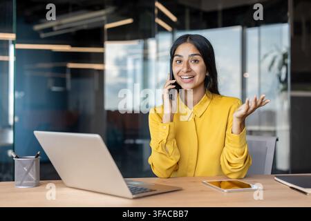 Giovane donna d'affari sorridente seduta alla scrivania in ufficio e al telefono. Foto Stock