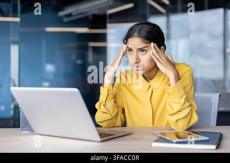 Giovane donna d'affari indiana stanca e sopraffatta seduta alla scrivania in ufficio e che tiene la testa. Foto Stock