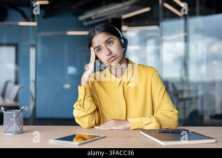 Giovane donna indiana stanca in abito seduta a una scrivania in ufficio e che tiene la testa. Foto Stock
