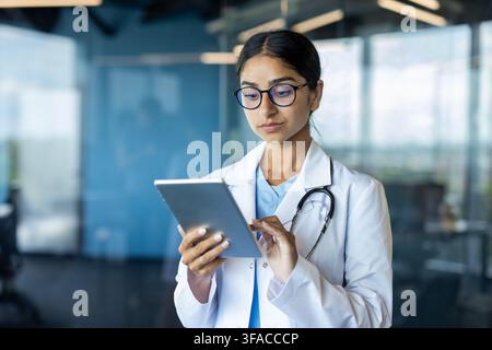 Giovane dottoressa indiana in camice bianco in piedi in una stanza della clinica e che usa un tablet. Foto Stock