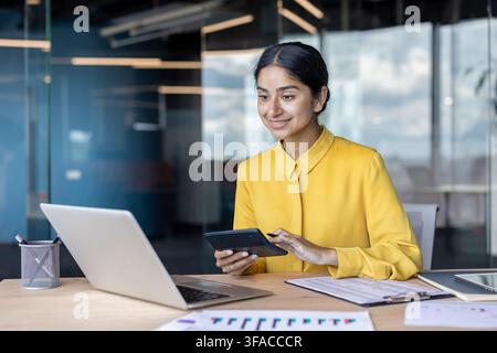Giovane contabile indiana seduta in ufficio, lavora con documenti e laptop, usa una calcolatrice. Foto Stock