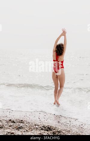 Donna in spiaggia, giovane donna in costume da bagno rosso che allunga le braccia mentre è in piedi su una spiaggia di ciottoli in riva al mare durante le vacanze estive Foto Stock