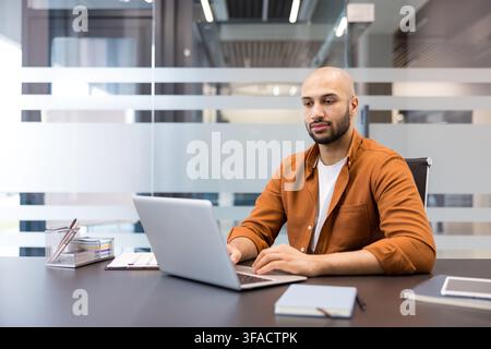 Giovane uomo con la barba concentrato sul suo laptop, che lavora diligentemente a una scrivania in un ambiente d'ufficio contemporaneo, rappresentando concetti di produttività, business e tecnologia Foto Stock
