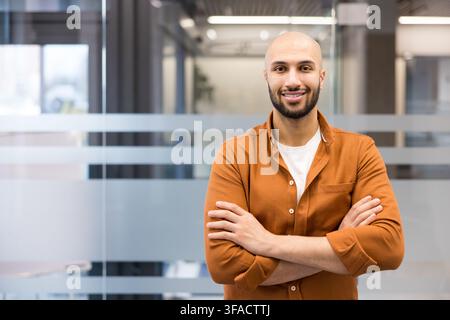Giovane uomo calvo e sicuro di sé con barba e maglietta bianca sotto una camicia color ruggine, in piedi a braccia incrociate e sorride alla fotocamera in un ambiente d'ufficio moderno Foto Stock