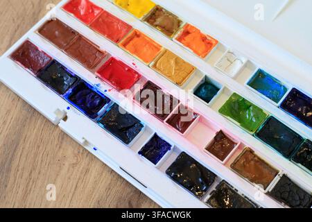 Panetti di pittura acquerello vivaci dentro una scatola in plastica bianca. Disposizione di un set artistico su superficie di legno in ambiente interno Foto Stock