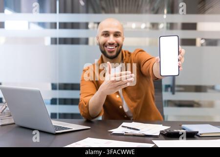 Uomo d'affari sorridente che tiene uno smartphone con uno schermo bianco vuoto, presentando una nuova app o servizio mentre è seduto a una scrivania con un laptop e documenti in un ufficio moderno Foto Stock
