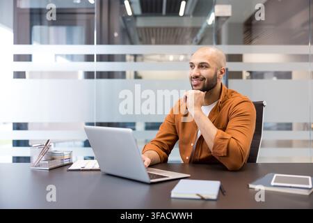 Giovane uomo d'affari calvo che sorride e riflette mentre lavora su un laptop a una scrivania contemporanea in ufficio, meditando su idee future e crescita professionale Foto Stock