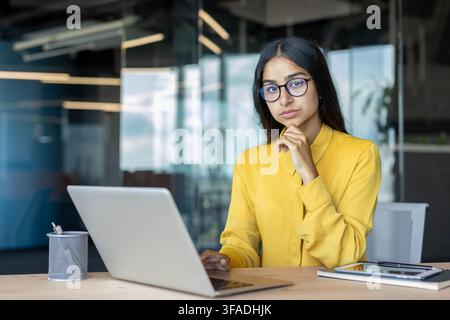 Ritratto di una giovane donna d'affari indiana seria con occhiali seduta a una scrivania con un laptop. Foto Stock