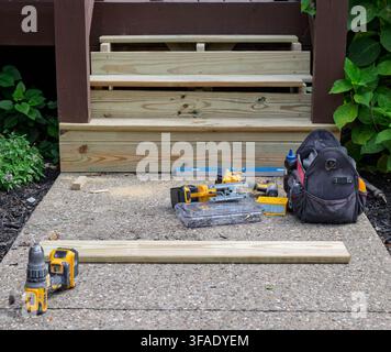 I gradini di ricambio del ponte in legno poggiano su un marciapiede di cemento. Gli attrezzi da costruzione sono a terra. Una borsa degli attrezzi riposa accanto alle assi. Piante verdi frondose crescono Foto Stock