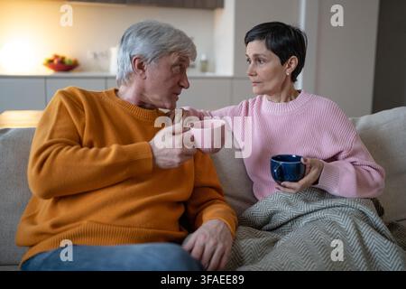 Moglie pensierosa che ascolta il marito anziano mentre la coppia sposata si rilassa insieme a casa sul divano. Foto Stock