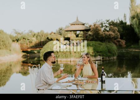 Una coppia si gode una cena intima a un tavolo elegante vicino all'acqua. Condividono risate e cibo raffinato, circondati dalla bellezza della natura. Foto Stock