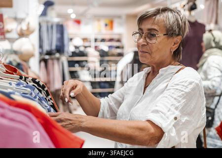 Donna caucasica anziana con capelli grigi corti che fa acquisti di abbigliamento in un negozio al dettaglio. Sta esaminando una camicia colorata mentre sfoglia uno scaffale di Foto Stock