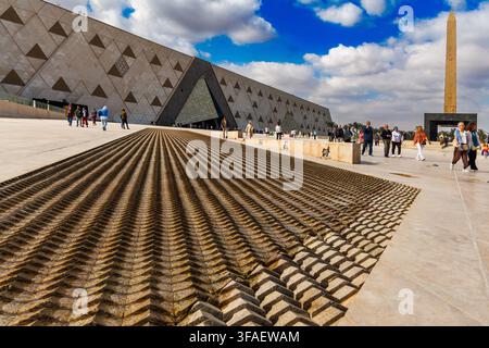 I turisti camminano attraverso la piazza davanti all'ingresso del Grande Museo Egiziano, con un antico obelisco sullo sfondo, Giza, Egitto. Foto Stock