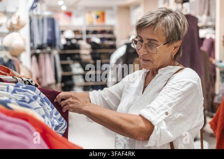 Donna caucasica anziana con capelli grigi corti che fa acquisti di abbigliamento in un negozio al dettaglio. Sta esaminando una camicia colorata mentre sfoglia uno scaffale di Foto Stock