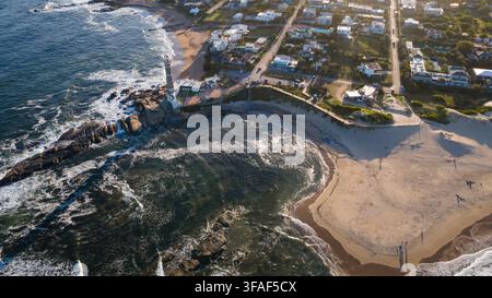 Scatto aereo che cattura il faro storico di José Ignacio su una penisola rocciosa, con onde che si infrangono contro la riva e una spiaggia sabbiosa sottostante. Il co Foto Stock