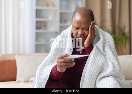 Uomo nero malato avvolto in una coperta a casa, che controlla la temperatura con un termometro Foto Stock