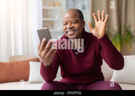 Uomo nero sorridente seduto a casa che saluta durante una videochiamata su smartphone Foto Stock
