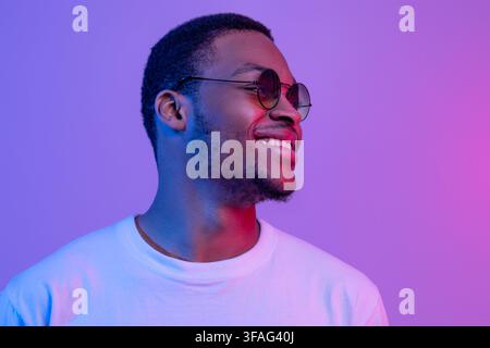 Sorridente e bello Black Millennial Guy in eleganti occhiali che si posano sotto Luce al neon Foto Stock