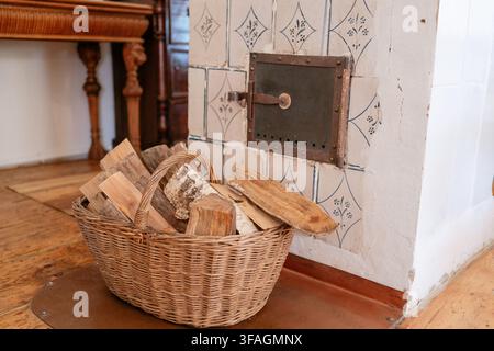 Un cesto di vimini pieno di legna da ardere si trova in un'accogliente area camino interna, che mostra un ambiente rustico e caldo con pavimento in legno e piastrelle in ceramica Foto Stock