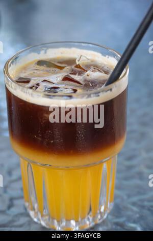 Un bicchiere rinfrescante di caffè freddo stratificato fatto con succo d'arancia brillante e caffè espresso scuro, con cubetti di ghiaccio e una cannuccia nera su una superficie testurizzata Foto Stock