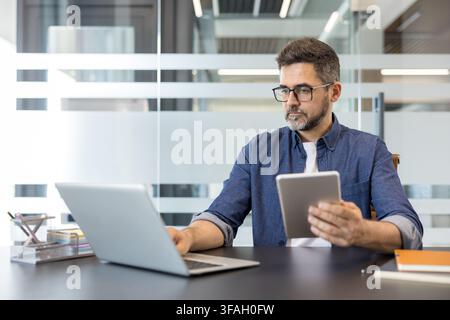 Uomo professionista concentrato sul lavoro, impegnato simultaneamente con un laptop e un tablet digitale, dimostrando efficienza e produttività moderna in un ambiente d'ufficio contemporaneo Foto Stock