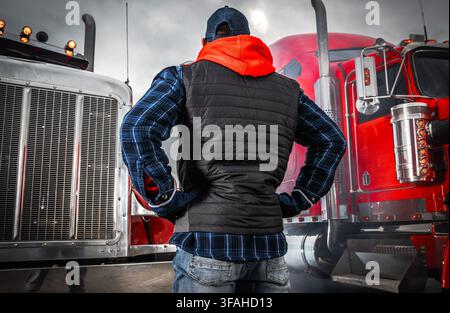 Un uomo in un gilet nero e camicia a quadri è in piedi vicino a un grande camion rosso in un deposito di camion. Il cielo è nuvoloso e la scena mostra un'atmosfera da cultura dei camionisti Foto Stock