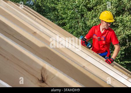 Un operaio che indossa un casco giallo e una camicia rossa sta utilizzando un utensile elettrico per installare travi di legno su un tetto. Il cantiere è circondato da alberi Foto Stock